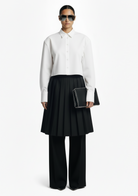 GOLD x TEAL Monarch Pleat Trousers shown on model in black wide-leg silhouette with detachable pleated skirt overlay, styled with a white shirt.