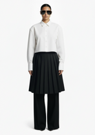 GOLD x TEAL Monarch Pleat Trousers shown on model in black wide-leg silhouette with detachable pleated skirt overlay, styled with a white shirt.